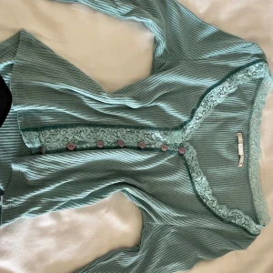 Blå/grön ribbad topp från Urban Outfitters - Supersöt blå/grön ribbad topp från Urban Outfitters med spetsdetaljer längs v-ringningen och knäppning framtill med lila knappar. Långärmad modell med extra spets vid ärmslut och längs knappslån. As gullig tröja som använts kanske 5 gånger som max. 
