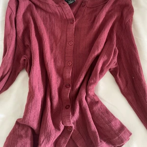 Vinröd kofta från Urban Outfitters - Vinröd kofta från Urban Outfitters i stickad bomull med snyggt hålmönster. Långärmad modell med knappar framtill och ribbade detaljer vid kanterna. Perfekt till hösten. Använt ca 2 gånger.