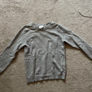 Grå stickad tröja från Vila - En enkel och stilren grå stickad tröja från Vila. Tröjan har rund halsringning, långa ärmar och ribbade muddar vid ärmslut och nederkant. Perfekt att matcha med jeans eller kjol för en clean look.