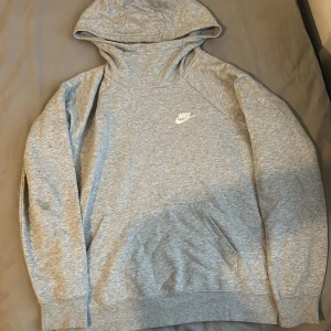Grå Nike hoodie med huva, storlek M - Snygg grå hoodie från Nike med klassisk logga broderad på bröstet. Tröjan har huva, känguruficka och långa ärmar. Perfekt för chill eller träning, i mjukt bomullsmaterial. Enkel och stilren design som funkar året runt.