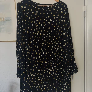Svart blommig långärmad klänning - Säljer en svart klänning med små gula och vita blommor. Klänningen har lång ärm och en rak, avslappnad passform. Perfekt för dig som gillar blommigt och vill ha en enkel men snygg look. Rund halsringning och mjukt, lätt material.