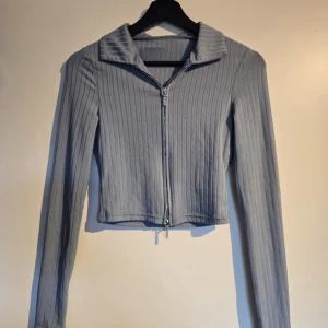 Ljusblå ribbad ziptop med krage - Säljer en ljusblå croppad topp med långa ärmar och dragkedja framtill. Toppen har en snygg krage och är ribbad för extra stilpoäng. Perfekt att matcha med jeans eller kjol för en trendig look.