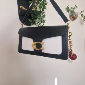 Coach svartvit axelväska i skinn - Coach axelväska i svart och vitt skinn med stilren rektangulär form. Väskan har en bred axelrem, guldiga metalldetaljer och ett stort C-spänne framtill. Utsidan är slät och insidan har dragkedja med Coach-logga. Dekorerad med pärlor och röd berlock. Berlocken följa inte med. (Köpte från shein) 