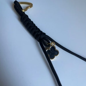 Svart nyckelring med gulddetaljer och berlock väska accessoarer  - Stilrent svart nyckelring med flätad design och guldfärgat hjärtlås. Nyckelringen har en liten fyrklöverformad berlock i svart och guld som ger en exklusiv touch. Perfekt accessoar för att lyfta din outfit.
