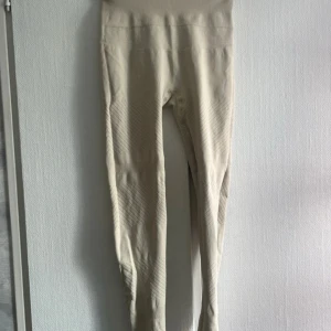 Tränings tights - Säljer ett par beige tränings tights från Casall med hög midja och diskret ribbade detaljer längs benen. De är stretchiga och sitter tight, perfekta för träning eller chill. Materialet är mjukt och följsamt syntet, vilket ger en skön passform.