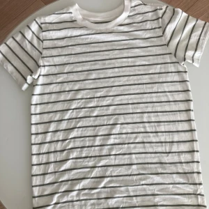 Vit randig t-shirt 100% Bomull  - Vit t-shirt från Kappahl med tunna horisontella svarta ränder. Klassisk rund halsringning och korta ärmar. Tillverkad i mjuk bomull, perfekt för en chill och stilren look.