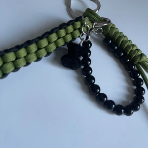 Grönt och svart armband med pärlor - Unikt armband med flätad grön och svart snodd, dekorerat med svarta pärlor och en liten svart björnberlock. Sitter på en metallring som gör det enkelt att fästa i byxan eller väskan. Perfekt accessoar för en cool och personlig stil.
