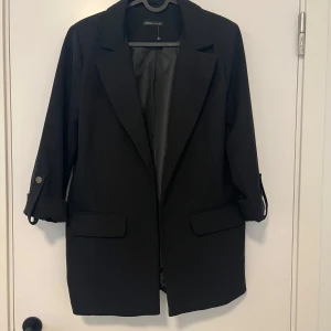 Svart kavaj från Divided, stl 38 - Stilren svart kavaj från Divided med klassisk krage, uppvikta ärmar med knappdetalj och två fickor framtill. Perfekt för en snygg och clean look. Tunn fodring på insidan och rak passform.