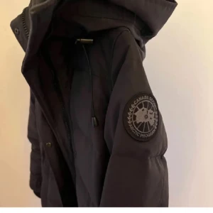 Svart dunjacka från Canada Goose - Säljer en svart, lång dunjacka från Canada Goose black label med huva och dragkedja. Jackan har en snygg patch på ärmen, stora fickor framtill och ribbade muddar vid ärmsluten. Perfekt för kalla dagar när du vill hålla stilen och värmen. 