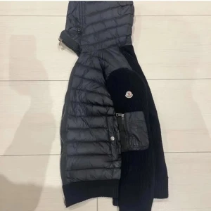 Svart Moncler Cardigan - Svart cardigan från Moncler med stickade ärmar. Jackan har huva, dragkedja framtill och en ficka med dragkedja på ärmen. Klassisk Moncler-logga på vänster ärm. Perfekt för kyliga dagar och riktigt snygg, inga skador. Skriv för funderingar