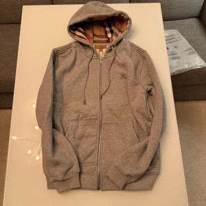 Grå Burberry hoodie med dragkedja - Snygg grå hoodie från Burberry med klassiskt rutigt foder i huvan och insidan. Tröjan har dragkedja framtill, snörning i huvan och två fickor. Tillverkad i mjuk bomullsmix och har ribbade muddar vid ärmslut och nederkant. Perfekt för en chill och stilren look.
