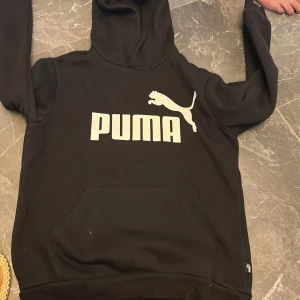 Svart hoodie från Puma med logga - Svart hoodie från Puma med stor vit logga och text på bröstet. Klassisk känguruficka framtill, ribbade muddar och huva. Perfekt för chill dagar eller när du vill ha en sportig vibe. Mjuk och skön i bomullsmix.
