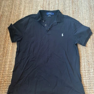 Svart pikétröja från Ralph Lauren - Klassisk svart pikétröja från Ralph Lauren i slim fit-modell med stretch mesh-material. Tröjan har korta ärmar, krage och knäppning framtill samt den ikoniska broderade loggan på bröstet. Perfekt för en clean och stilren look.