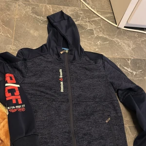Reebok CrossFit hoodie XL marinblå - Säljer en marinblå Reebok CrossFit hoodie i storlek XL. Tröjan har hel dragkedja, huva och tryck på bröstet och ärmen. Materialet är mjukt och funktionsanpassat, perfekt för träning. Snygga detaljer och sportig look.