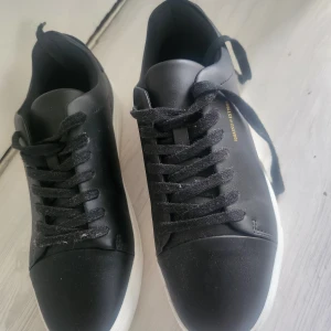 Svarta sneakers från Iguassu - Svarta sneakers från Iguassu med stilren design och vit platt sula. Skorna har snörning och är tillverkade i syntetmaterial. På sidan finns en guldfärgad textdetalj med 'IGUASSU - EST/1989'. Perfekta för en clean och modern look.