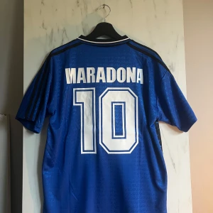 Argentina Maradona #10 blå fotbollströja - Säljer en ikonisk blå Argentina fotbollströja från Adidas med Maradona och nummer 10 på ryggen. Tröjan har korta ärmar, svart krage med vit kant, broderat landslagsmärke och ett grafiskt mönster i svart längs sidan. Materialet är glansigt polyester och trycket är i vitt.