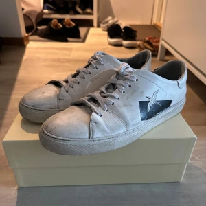 Vit sneaker från Axel Arigato - Snygga vita sneakers från Axel Arigato med klassisk låg profil och rund tå. Skorna har snörning och är tillverkade i skinn. På sidan finns en svart detalj med en broderad fågel. Tvättas såklart innan dom skickas.