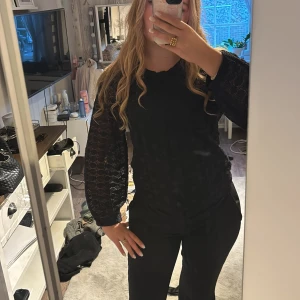 Svart spetsblus från Hallys - Säljer en svart blus från hallys med genombrutna spetsärmar och diskret blommönster i tyget. Blusen har rund halsringning och långa ärmar med resår vid ärmslut. Perfekt för dig som gillar en stilren men ändå detaljrik look.