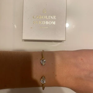 Guldarmband med kristaller Caroline Svedbom - Snyggt guldarmband från Caroline Svedbom med två små, droppformade klara kristaller i varje ände. Armbandet är öppet och har en tunn, elegant design som ger en lyxig vibe. Perfekt för dig som gillar bling och vill sticka ut med något glittrigt. Insidan har tappt lite färg men inget som syns när man bär det 