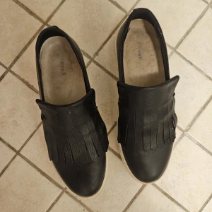 Svarta sneakers med fransar Filippa K - Svarta sneakers från Filippa K i skinn med coola fransdetaljer på ovansidan och vit platt sula. Snygg minimalistisk design med rund tå och slip-on-modell. Perfekta för dig som gillar stilrena men ändå unika skor.