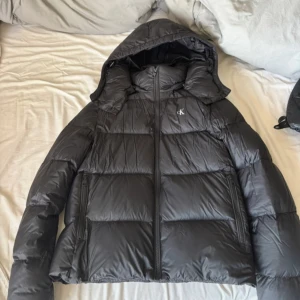 Svart pufferjacka Calvin Klein Jeans - Svart pufferjacka från Calvin Klein Jeans med huva och dragkedja framtill. Jackan har quiltad design, två sidofickor med dragkedja och logga på bröstet samt ärmen. Perfekt för kalla dagar och har en modern, lite boxig passform. Ändå defekten är att texten på armen har försvunnit lite men inget man tänker på💞