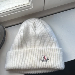 Vit ribbad mössa från Moncler - Snygg vit mössa från Moncler med ribbad stickning och uppvikt kant. Klassisk Moncler-logga framtill och mjukt material som håller dig varm. Perfekt för dig som vill ha en clean och exklusiv look under kalla dagar. Man kommer även in på monclers sida när man scannar den