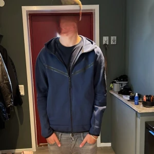 Mörkblå nike tech hoodie - Snygg mörkblå hoodie med hel dragkedja och huva. Tröjan har långa ärmar och diskreta detaljer på bröstet. Perfekt för chill dagar och enkel att matcha med jeans eller joggers. Materialet känns mjukt och bekvämt mot huden.