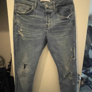 Blå slitna jeans från Zapa - Säljer ett par blå jeans från Zara med snygga slitningar och hål på benen. Jeansen har klassisk femficksdesign, normal midja och smal passform. Perfekta för dig som gillar en avslappnad och trendig look.