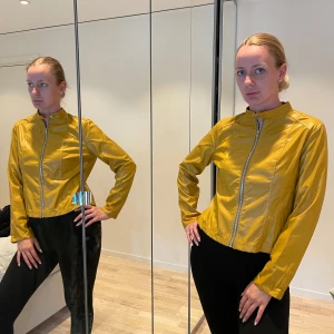 Gul skinnjacka med dragkedja - Snygg gul jacka med rak passform och hög krage med tryckknapp. Jackan har en silvrig dragkedja framtill och långa ärmar. Perfekt för dig som vill sticka ut med en cool färg och stilren look.