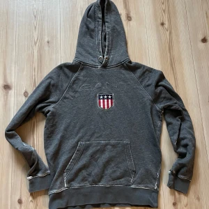 Grå hoodie från GANT - strl.M - Mörkgrå hoodie från GANT med broderad logga och amerikanskt inspirerat emblem på bröstet. Tröjan har huva med snörningshål, magficka och ribbade muddar. Perfekt för dig som gillar klassisk och sportig stil.
