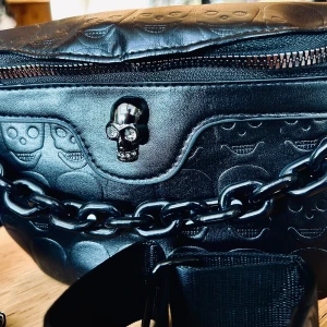 Rockig väska i PU-läder med dödskallar från Scull Cult! - Mångasidig  svart rockig väska i PU-läder med dödskallar från Scull Cult som kan bäras som midjeväska, crossbodybag eller axelväska. Snygga detaljer och ett stort och ett litet fack som stängs med dragkedja och justerbar axelrem Unisexmodell. Mått: 20 x 15 cm. Oanvänd.
