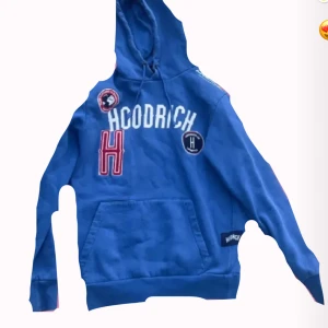 Blå hoodie från Hoodrich XS - Blå hoodie från Hoodrich med broderade patches och stor vit logga på bröstet. Tröjan har huva med snörning och en stor magficka. Snygga röda och vita detaljer ger en sportig vibe. Perfekt för dig som gillar streetwear.
