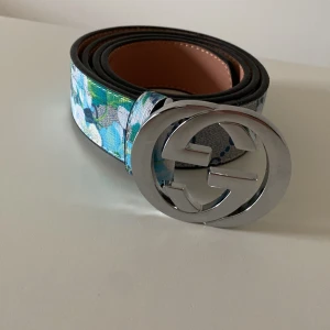G blommigt bälte med silver GG-spänne - Snyggt bälte från GGmed stort silverfärgat GG-spänne. Bältet har ett blommigt mönster i blått, grönt och vitt på klassisk GG-canvas. Insidan är i ljust skinn. Perfekt accessoar för att lyfta din outfit.