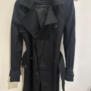 Svart trenchcoat med bälte från H&M - Stilren svart trenchcoat från H&M med dubbelknäppning, klassisk krage och bälte i midjan. Jackan har långa ärmar med spänndetaljer vid ärmsluten och stora knappar framtill. Perfekt för dig som vill ha en tidlös och snygg look.