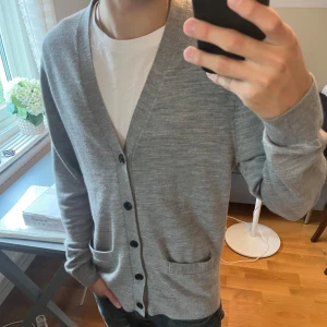 Grå cardigan från Abercrombie & Fitch - Snygg grå kofta från Abercrombie & Fitch i klassisk modell med v-ringning och svarta knappar. Koftan har två fickor framtill och är tillverkad i mjukt material som passar perfekt till lager-på-lager. Enkel att matcha med både t-shirt och skjorta.