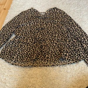 Leopardmönstrad blus från Kappahl - Snygg blus från Kappahl i leopardmönster med volangdetalj i midjan. Toppen har långa ärmar och är i beige och svart. Perfekt för dig som vill sticka ut lite extra med ett djurmönster.