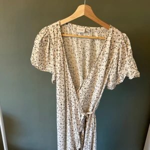 Blommig omlottklänning från Vila - Supersöt omlottklänning från Vila i ljusbeige med små blommor i rosa och grönt. Klänningen har korta puffärmar, v-ringning och knyt i midjan. Perfekt för varma dagar och ger en romantisk vibe.
