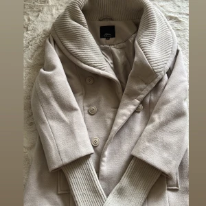 Beige kappa  - Snygg beige kappa med dubbelknäppning och bred ribbad krage. Jackan har ribbade muddar vid ärmsluten och två fickor framtill. Perfekt för dig som vill ha en stilren och mysig jacka med coola detaljer.