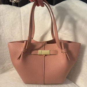 Rosa väska från Lindex  - Snygg rosa axelväska i skinnimitation med justerbar axelrem och två handtag. Väskan har en guldfärgad detalj framtill som ger en chic touch. Insidan har en praktisk ficka med dragkedja och magnetlås. Perfekt storlek för det viktigaste och stilren design. Säljs inte längre hos Lindex där med priset.