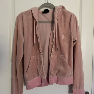 Rosa velour hoodie från Juicy Couture - Supermjuk rosa hoodie från Juicy Couture i velour med dragkedja, huva och snörning. Klassisk logga broderad på bröstet och två fickor framtill. Perfekt för en chill och trendig look. Storlek S. Passar XS. 