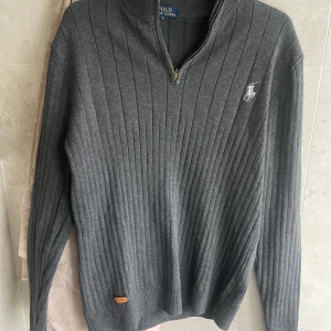 Grå ribbad tröja från Polo Ralph Lauren - Snygg grå ribbstickad tröja från Polo Ralph Lauren med halv dragkedja och broderad logga på bröstet. Tröjan har hög krage och långa ärmar, perfekt för lager-på-lager under kyliga dagar.