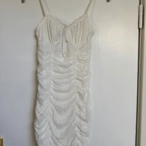 Vit omlott kortklänning med spets - Supersöt vit kortklänning från H&M med smala axelband och omlott-detalj vid bysten. Klänningen har snygg rynkning längs hela framsidan och är gjord i ett spetsliknande material som ger en romantisk vibe. Perfekt för sommarens alla tillfällen!