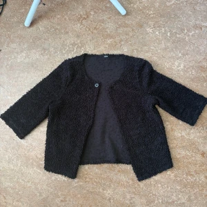 Svart fluffig kofta med knapp - Mysig svart kofta med fluffig textur och en enkel knapp framtill. Koftan har trekvartsärmar och är perfekt för lager-på-lager-stil. Den har en rak passform och känns både trendig och bekväm, passar till många olika outfits.