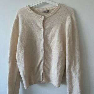 Beige kofta i ullblandning från H&M - Mysig beige kofta från H&M i ullblandning med rund halsringning och knappar framtill. Långärmad modell med ribbade muddar vid ärmslut och nederkant. Perfekt för lager-på-lager under kyliga dagar.