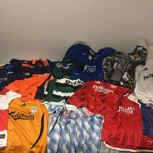 Samling av fotbollströjor från toppklubbar - Fet samling fotbollströjor från klubbar som Arsenal, Liverpool, Barcelona, Manchester United, Sporting, Chelsea och fler. Tröjorna är i olika färger som röd, blå, grön, orange, vit och guld, med klassiska klubbmärken, tryck och mönster som ränder, blixtar och rutigt. Perfekt för dig som älskar fotboll och vill ha unika matchtröjor.
