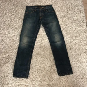 Mörkblå Nudie Jeans 31/34 straight fit - Säljer ett par mörkblå jeans från Nudie Jeans i modellen straight fit. Klassisk femficksdesign, orangea sömmar och snygga slitningar på låren. Jeansen är tillverkade i bomull och har Nudie-loggan på bakfickan.