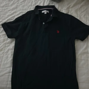 Mörkblå pikétröja från US Polo Assn - Mörkblå pikétröja från US Polo Assn med klassisk krage och knappslå. Röd broderad logga på bröstet ger en clean look. Tillverkad i mjuk bomull och har korta ärmar. Perfekt för en stilren och avslappnad outfit.