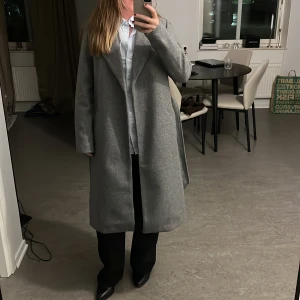 Grå kappa med bälte och slag - Snygg grå kappa med klassiska slag och knytskärp i midjan. Lång modell med raka ärmar och diskreta fickor på sidorna. Perfekt för dig som gillar stilrena och minimalistiska plagg. Kappan har en mjuk känsla och passar till många outfits. Oanvänd med lappen kvar, från vero moda 