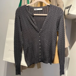 Mörkgrå kabelstickad kofta från Zara - Supersnygg mörkgrå kabelstickad kofta från Zara med glittriga knappar framtill. Koftan har lång ärm, v-ringning och är gjord i ett mjukt stickat material. Perfekt att slänga över en topp för en chill men ändå stilsäker look.