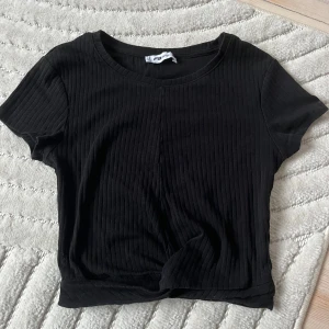 Svart ribbad topp med knyt  - Svart ribbad topp med korta ärmar och rund halsringning. Toppen har en croppad passform och en snygg knytdetalj framtill. Passar även S
